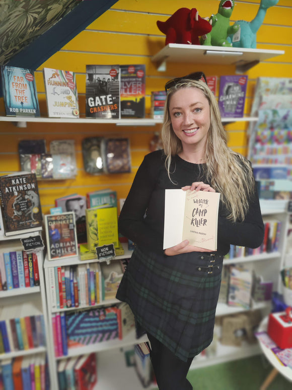 Cynthia Murphy - white female author holding a signed copy of her book Welcome To Camp Killer in front of book shelves in book shop B For Butterfly Books